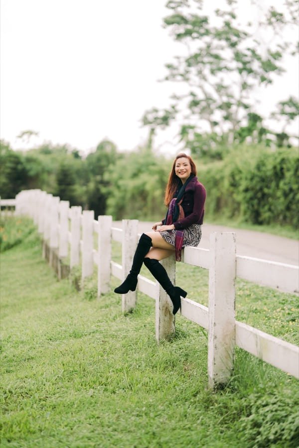Elyne Villamor seated on a white fence rail, in a plum cardigan and plaid scarf