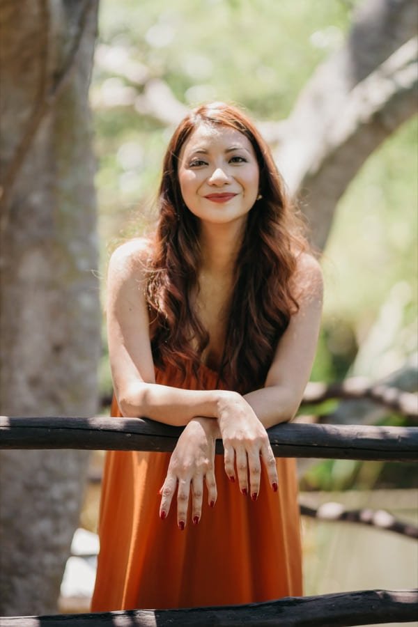 Portrait of Elyne Villamor in a rust dress, leaning on a wooden rail outdoors